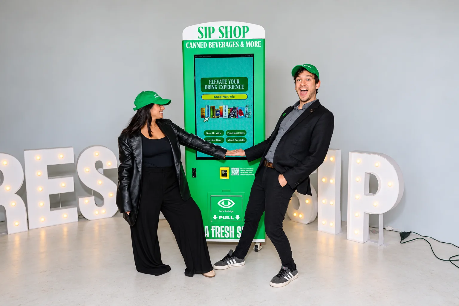 Two people in A Fresh Sip caps posing by the green Sip Shop smart vending kiosk at a launch event